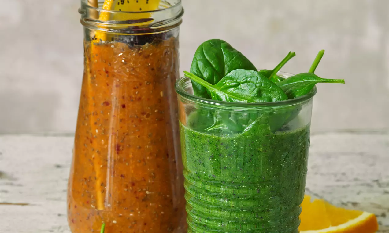 Zelené smoothie v pohári so štruktúrovaným povrchom zdobené čerstvými listami špenátu vedľa oranžového smoothie s plátkom pomaranča na vrchu