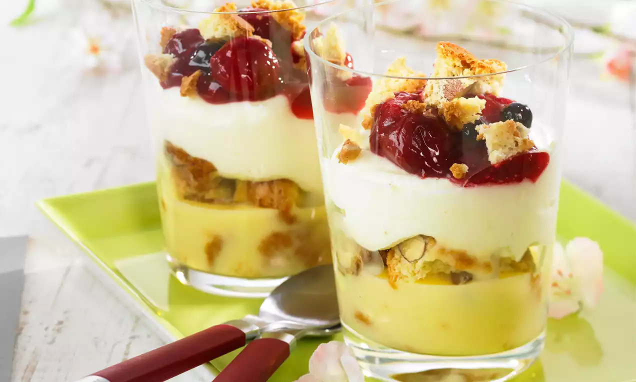 Schichtdessert in Gläsern mit gelber Creme, weißer Mascarponecreme, roten Fruchtstücken und zerbröselten Cantuccini auf grünem Teller