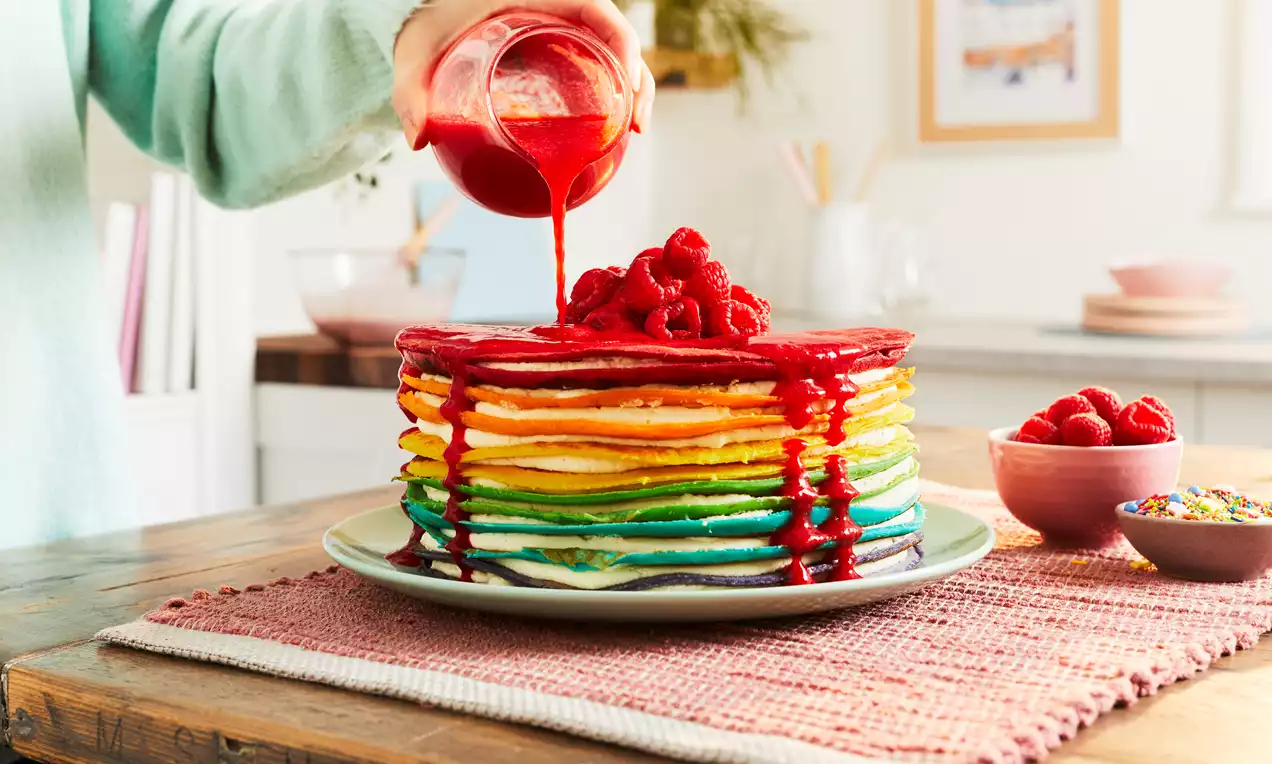 A stacked rainbow crepe cake with smooth layers in vibrant colours, topped with fresh raspberries and red sauce on a white plate