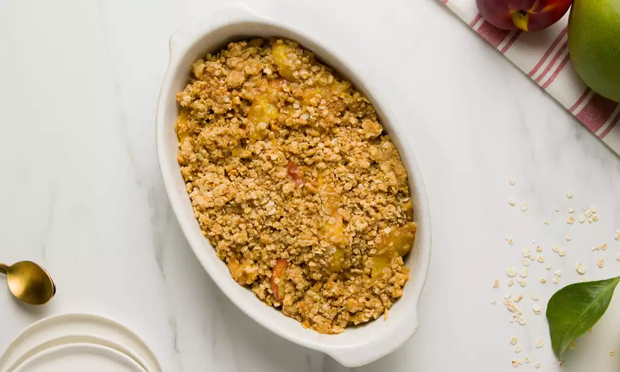 Crumble dorado con textura granulada sobre trozos de nectarinas y mango en una fuente blanca ovalada sobre mesa de mármol