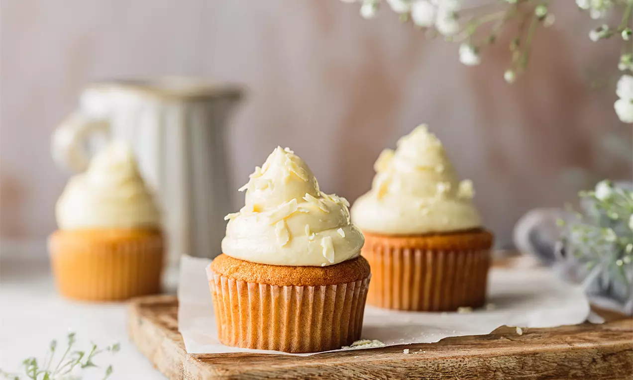 Picture - Cupcake dorati con soffice crema alla vaniglia color avorio decorati con scaglie di cioccolato bianco su un tagliere di legno