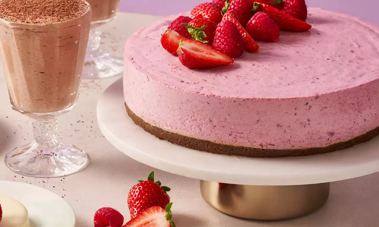 Ronde vegan taart met een roze romige vulling op een donkere bodem, gegarneerd met rood fruit en verse aardbeien op een wit taartplateau