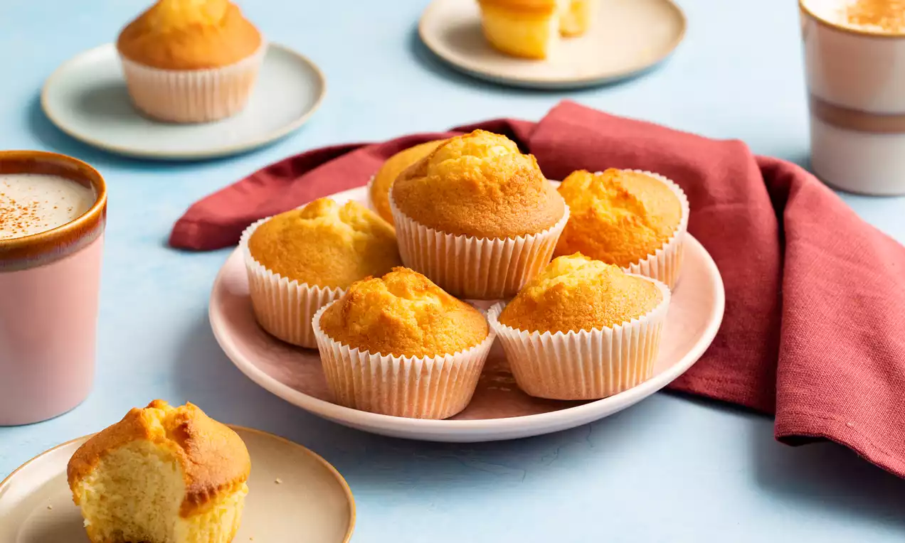 Goudgele, luchtige muffins in witte vormpjes op een roze bord met een rode servet, omringd door koffiekopjes en bordjes op blauw tafelblad