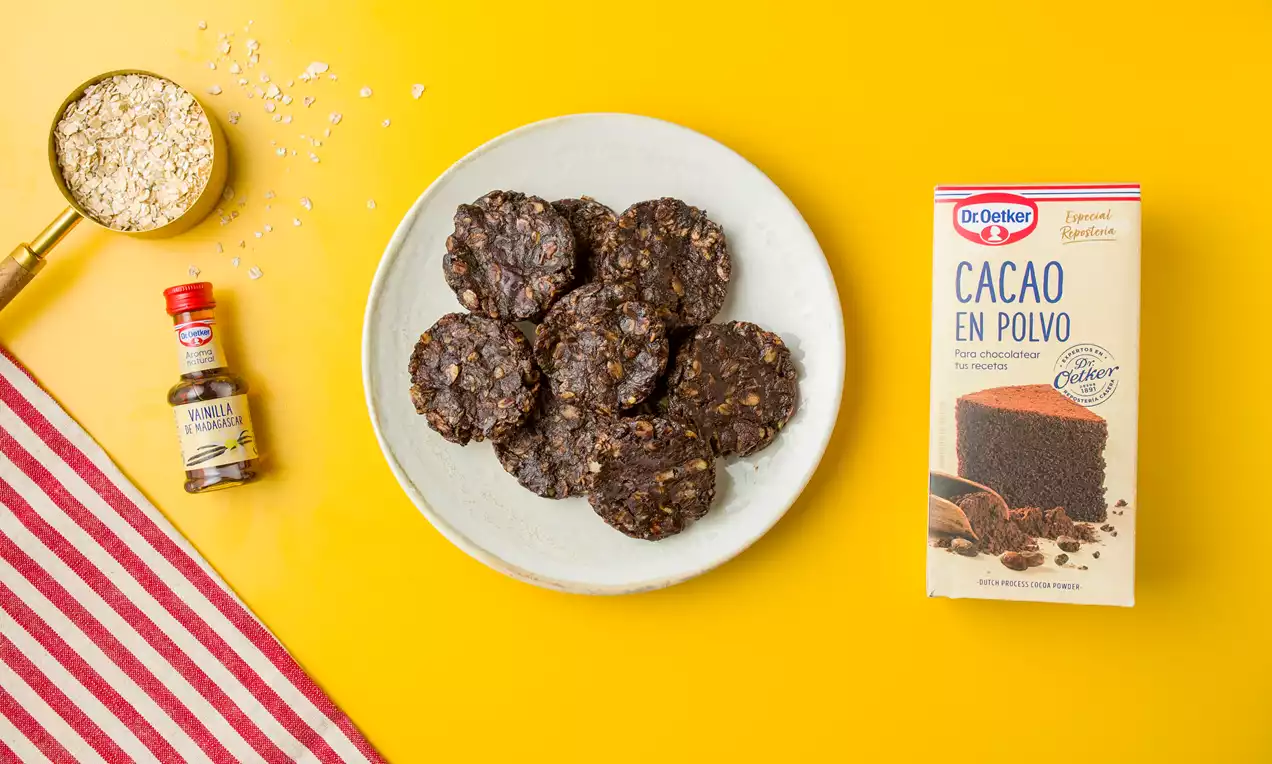 Galletas redondeadas de cacao oscuro con textura rugosa y copos de avena sobre plato blanco en mesa amarilla junto a ingredientes