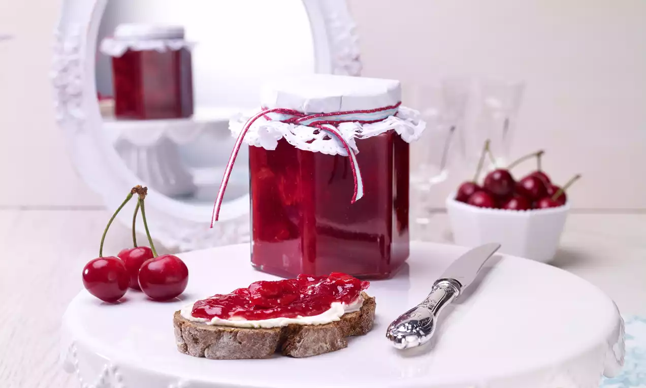 Confiture rouge brillante dans un pot décoré de dentelle, tranche de pain tartinée, cerises éclatantes et couteau argenté sur un présentoir blanc