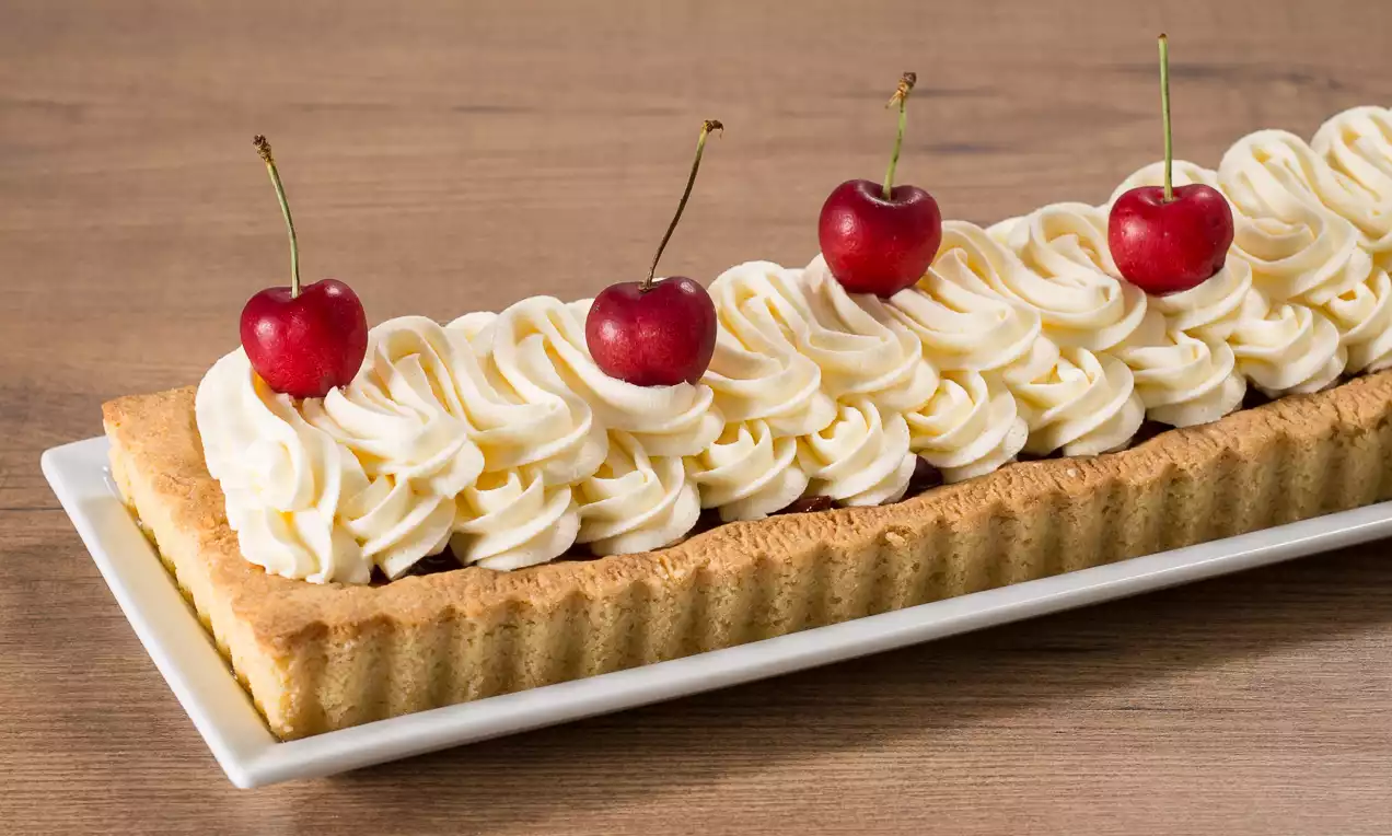 Tart rettangolare con bordo dorato, decorata con riccioli di crema bianca soffice e quattro ciliegie rosso vivo su un piatto bianco