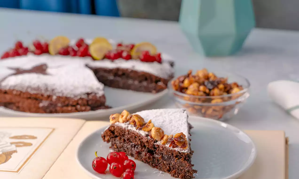 Felia de tartă vegană cu hrișcă, presărată cu zahăr pudră, decorată cu alune crocante și coacăze roșii pe o farfurie albă