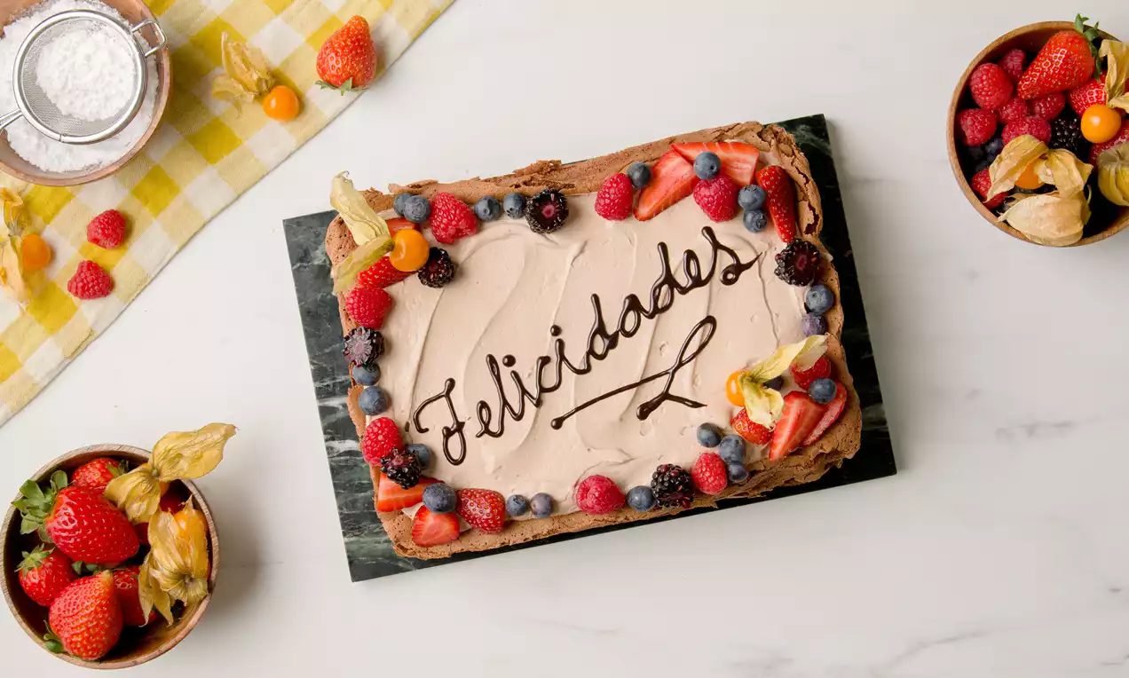 Tarta rectangular de chocolate con crema marrón clara, mensaje "Felicidades" en chocolate oscuro y bordes decorados con frutas frescas