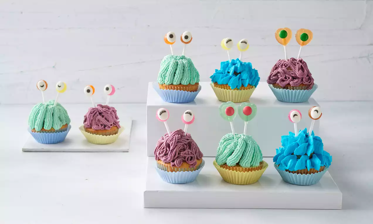 Monster-Cupcakes mit grüner, blauer und lilafarbener Creme, verziert mit bunten Zuckeraugen auf weißen Kuchenständern und Tellern platziert