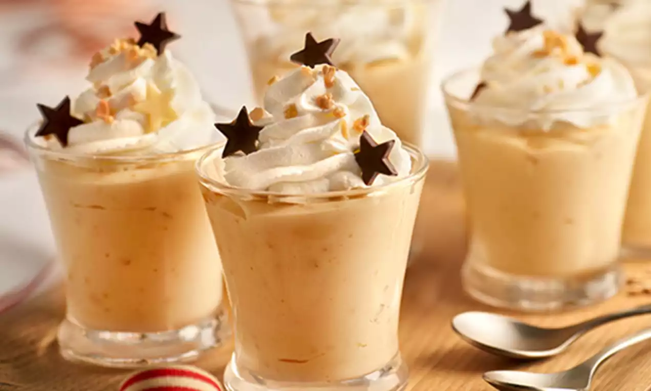 Vasos de mousse beige cremosa coronada con rosetones de nata, crocanti dorado y estrellas de chocolate negro sobre una tabla de madera