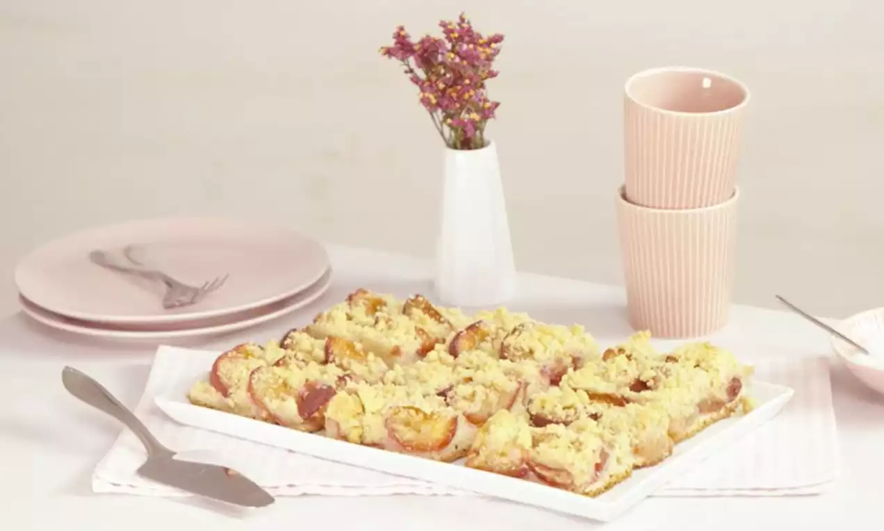 Pflaumen-Streusel-Kuchen mit goldgelbem Teig und buttrigen Streuseln, gebacken auf einem weißen Rechteck-Teller, neben rosa Geschirr