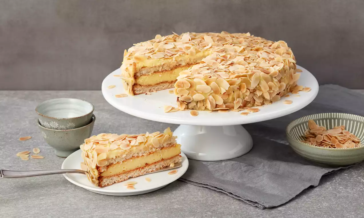 Schwedische Mandeltorte mit goldbrauner Mandelkruste, luftiger Vanillecreme-Schicht und Puderzucker, auf weißem Tortenständer angeschnitten