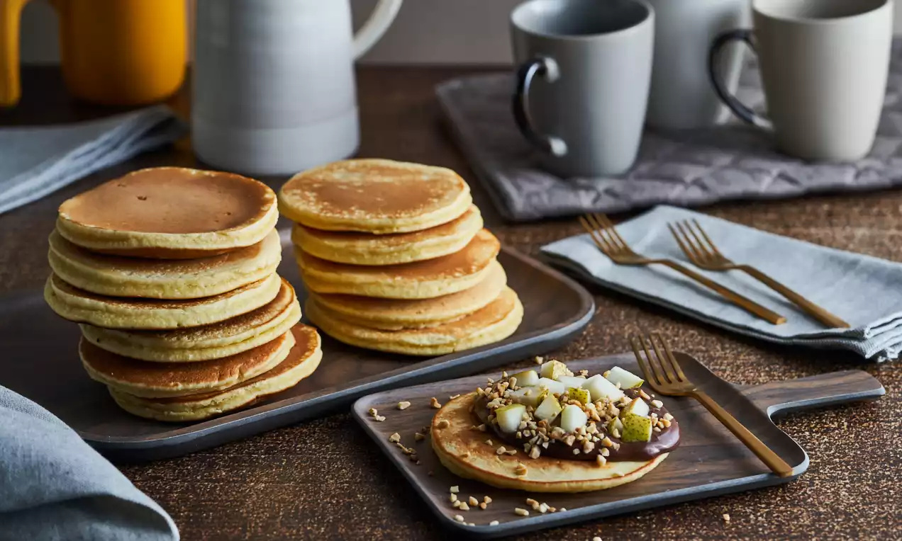 Pancakes dorati e soffici disposti in pile su un vassoio, uno decorato con cioccolato lucido, dadini di pera e granella croccante su un tagliere scuro
