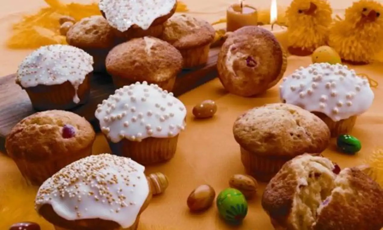 Muffins med gyldenbrun overflate, noen glacert og pyntet med perler, på gul duk med påskeegg, lys og påskekyllinger rundt