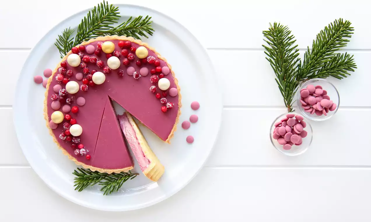 Ruby-terte med glatt rosa fyll, pyntet med hvite og røde sjokoladekuler og bær, på et hvitt fat dekorert med grønne grankvister