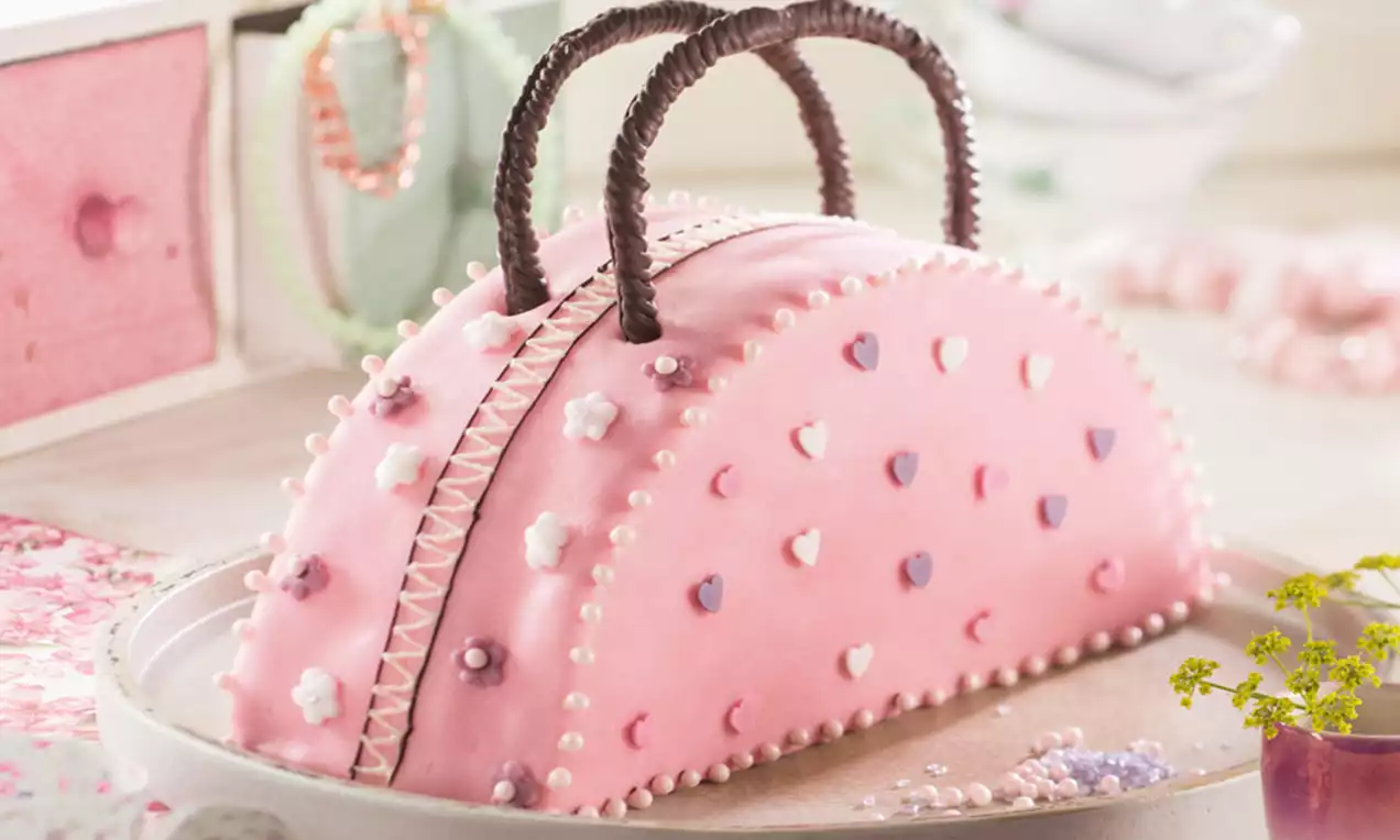 Gebogene rosafarbene Torte in Handtaschenform, verziert mit Perlen, Blüten und dunklen Griffen auf einem runden Tablett mit zarten Deko-Elementen