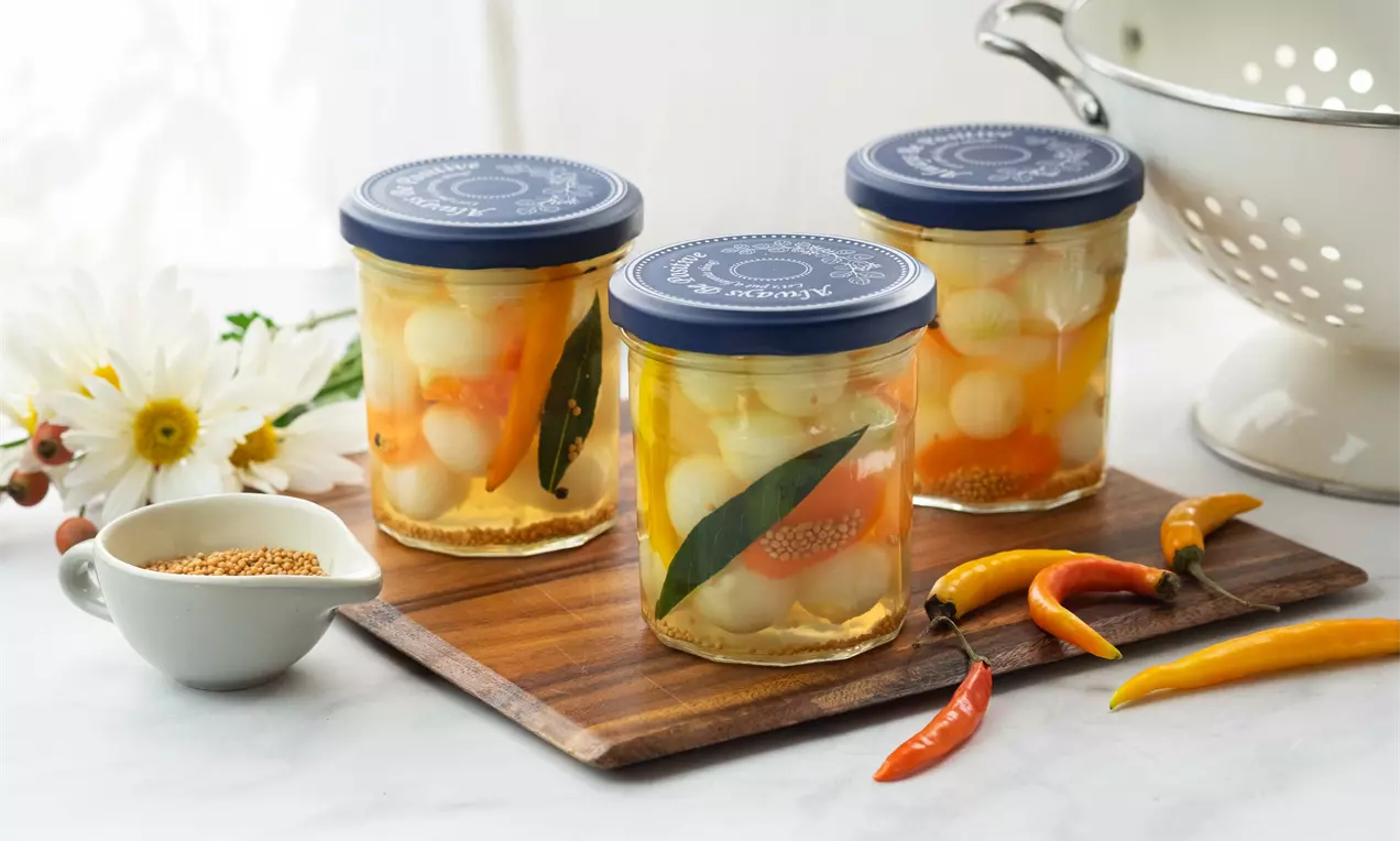 Перлени лукчета с гладка бяла повърхност, потопени в златиста марината с дафинов лист и ярки чушки върху дървена поставка