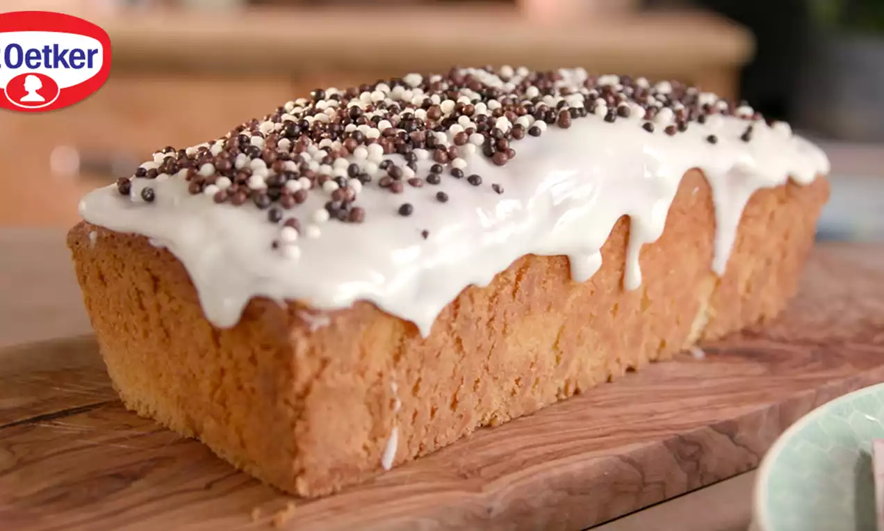 Cake doré rectangulaire avec un glaçage blanc crémeux coulant, parsemé de billes croustillantes chocolatées sur une planche en bois