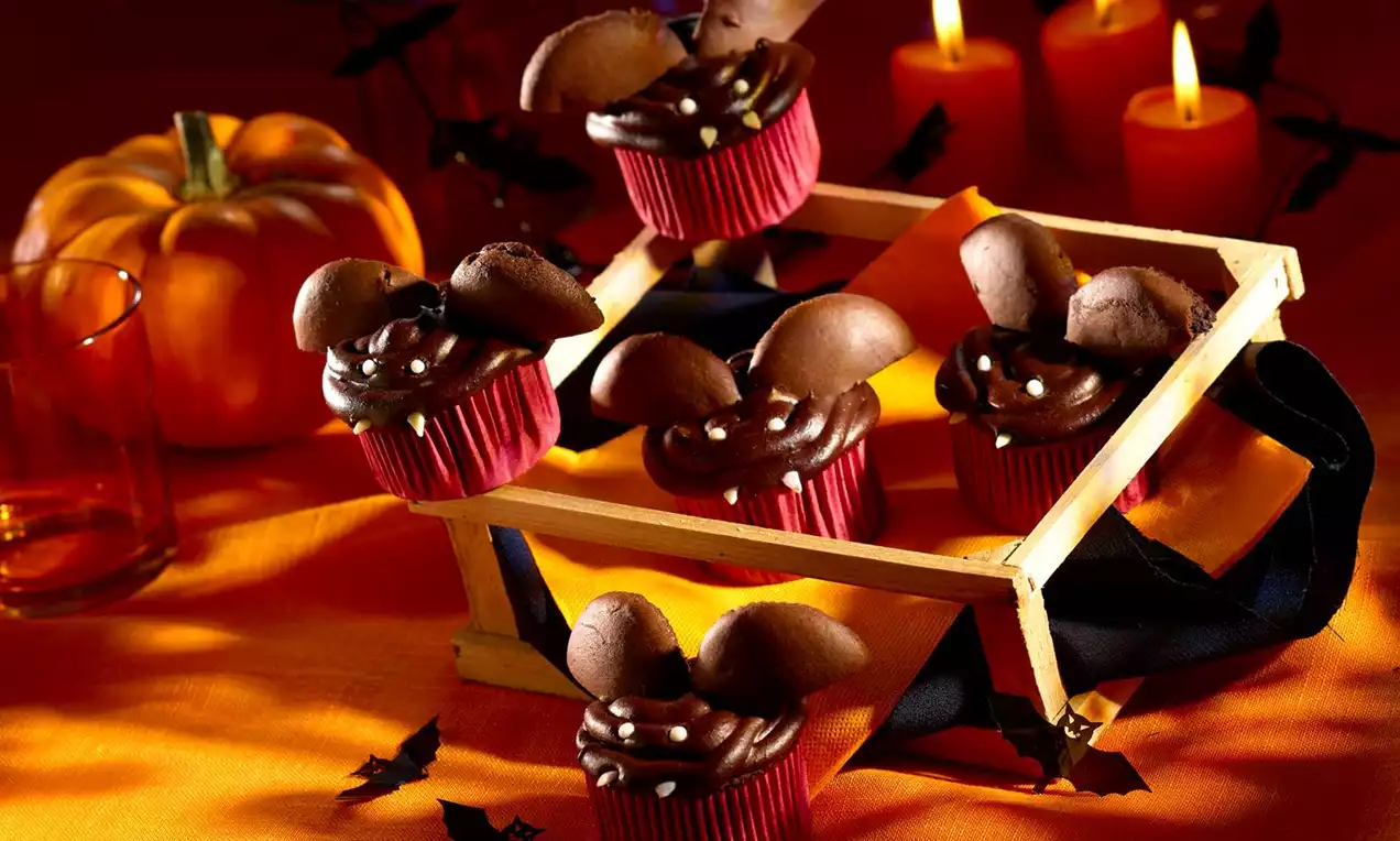 Cupcakes au chocolat foncé avec des oreilles en biscuit brun, décorés de perles blanches sur des caissettes rouges dans un décor halloween orange