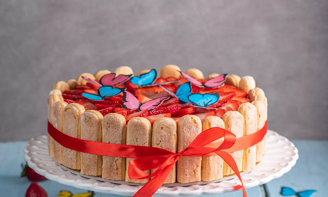Torta Šarlota s kremno sredico in oblogo iz svetlih piškotov, ovita z rdečim trakom, okrašena z jagodami in pisanimi metulji na beli stojali