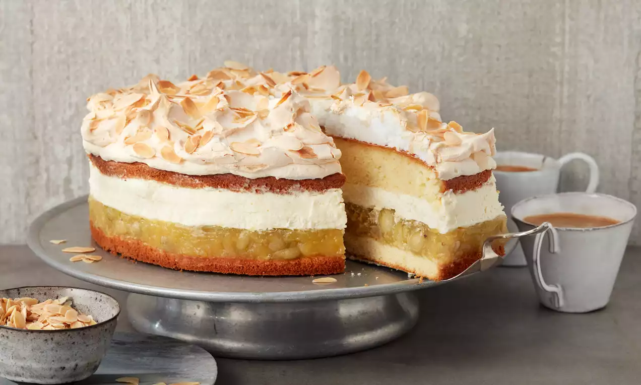 Stachelbeerkuchen mit goldbraunem Baiser und Mandelblättchen, geschichtet auf silberner Tortenplatte, daneben Schalen und Tassen aus Keramik
