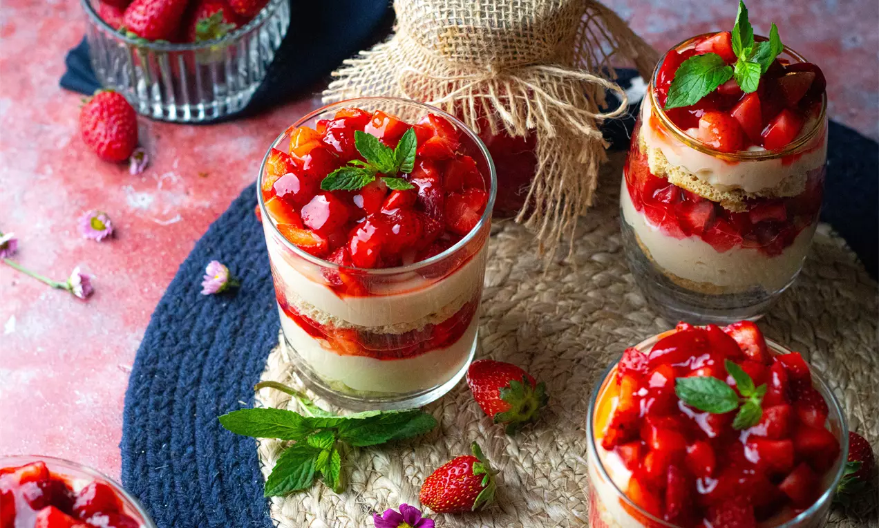 Баварска торта в прозрачна чашка с кремообразен светложълт слой, хрупкав бисквитен слой и яркочервен ягодов топинг, декорирана с листа мента