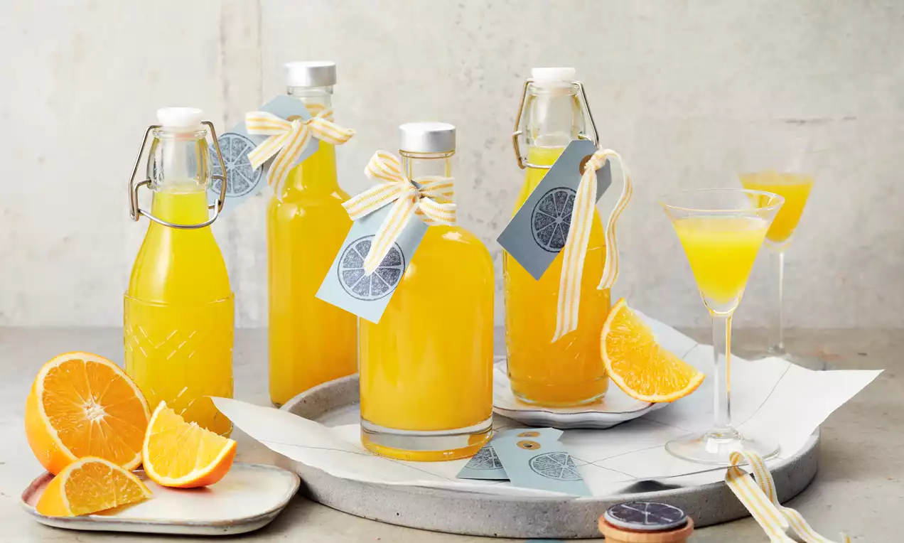 Leuchtend orangefarbener Likör in Glasflaschen mit weiß-gelber Schleife und Etikett, arrangiert mit frischen Orangenscheiben auf hellem Untergrund