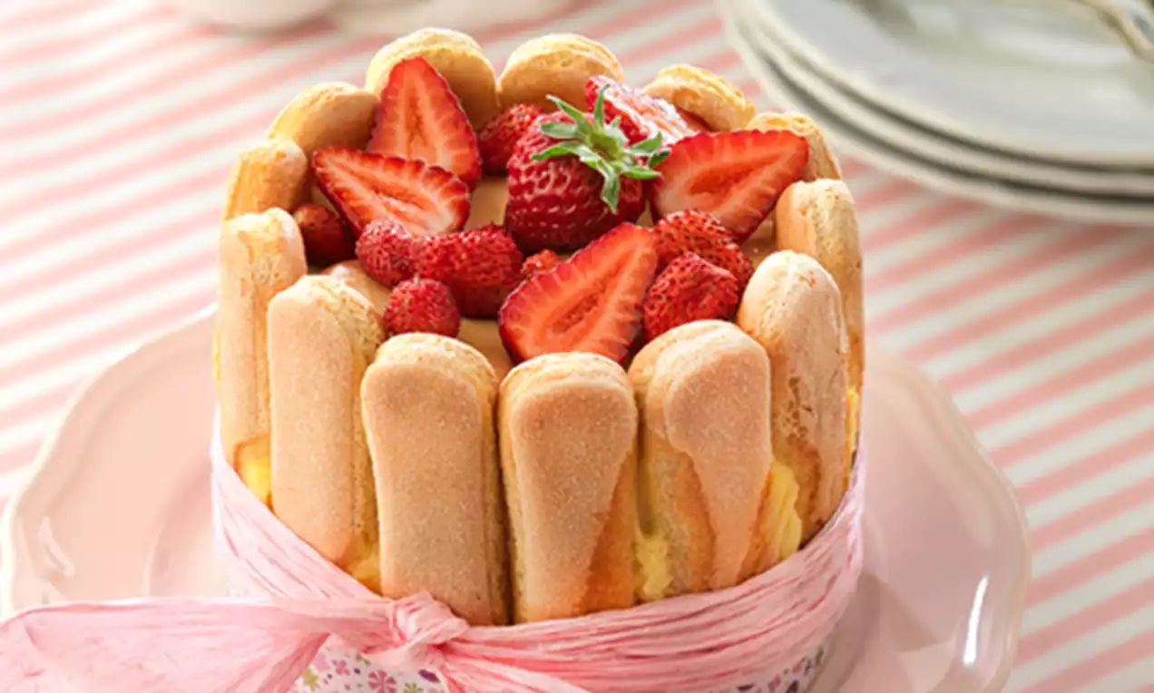 Charlota con bordes de bizcochos esponjosos verticales, coronada con fresas rojas brillantes sobre crema suave, rodeada por cinta rosa