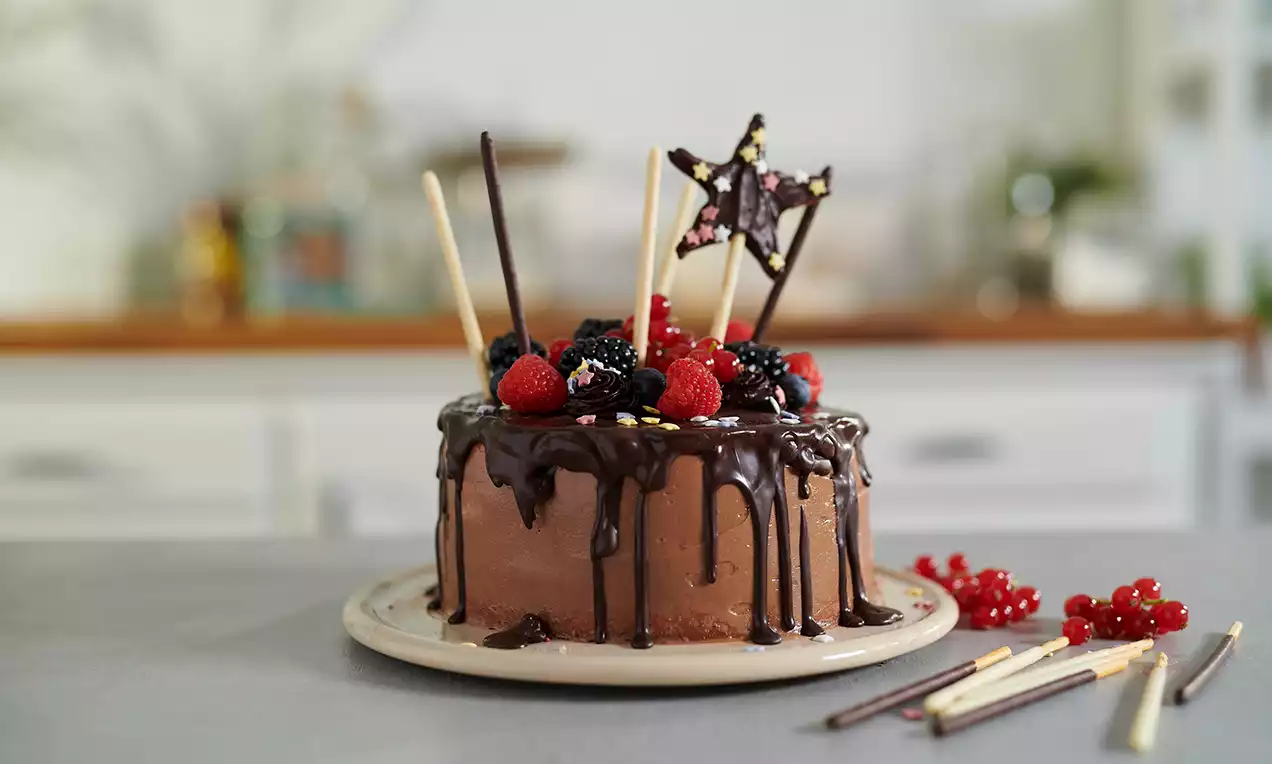 Gâteau rond au chocolat glacé de ganache brillante, orné de fruits rouges, bâtonnets colorés et étoile en chocolat sur une assiette beige