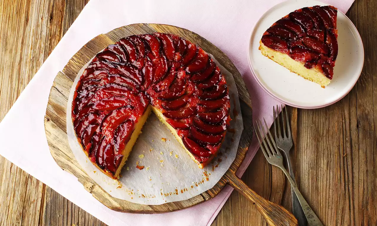 Tartın altın renkli yumuşak keki üzerinde dilimlenmiş parlak mor erikler spiral bir desenle sıralanmış, ahşap bir servis üzerinde kesilmiş