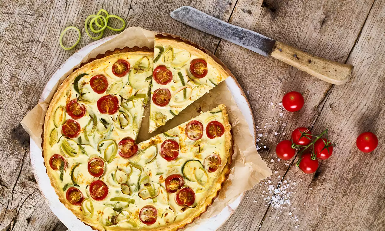 Goldbraune Gemüse-Quiche mit roten Tomatenhälften und grünen Streifen auf knusprigem Teigboden, serviert auf Backpapier und Holzuntergrund