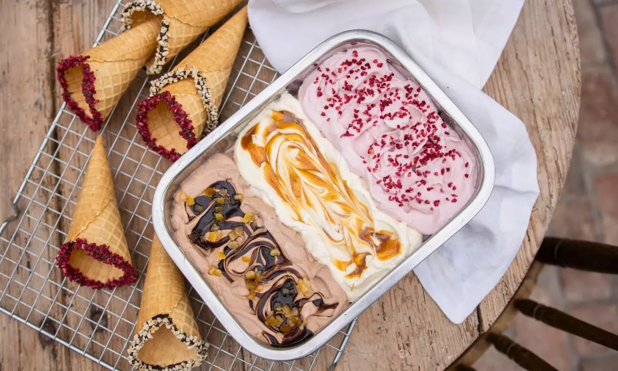 Metallform med tre sommarglassorter i rosa, vit och brun färg, toppat med bär, chokladsås och kolasås, bredvid glasstrutar på trådställ