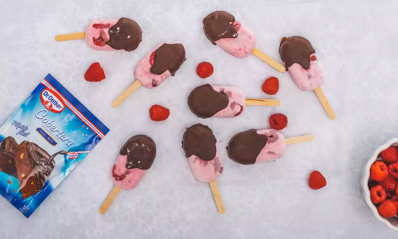 Polos rosados de frambuesa con cobertura de chocolate brillante, sobre palitos de madera, dispersos con frambuesas frescas en una superficie blanca