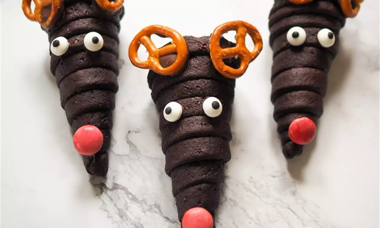 Три шоколадови конусчета с тъмнокафява текстура, декорирани с бели очи, червени муцунки и солени бретцел уши върху мраморен фон