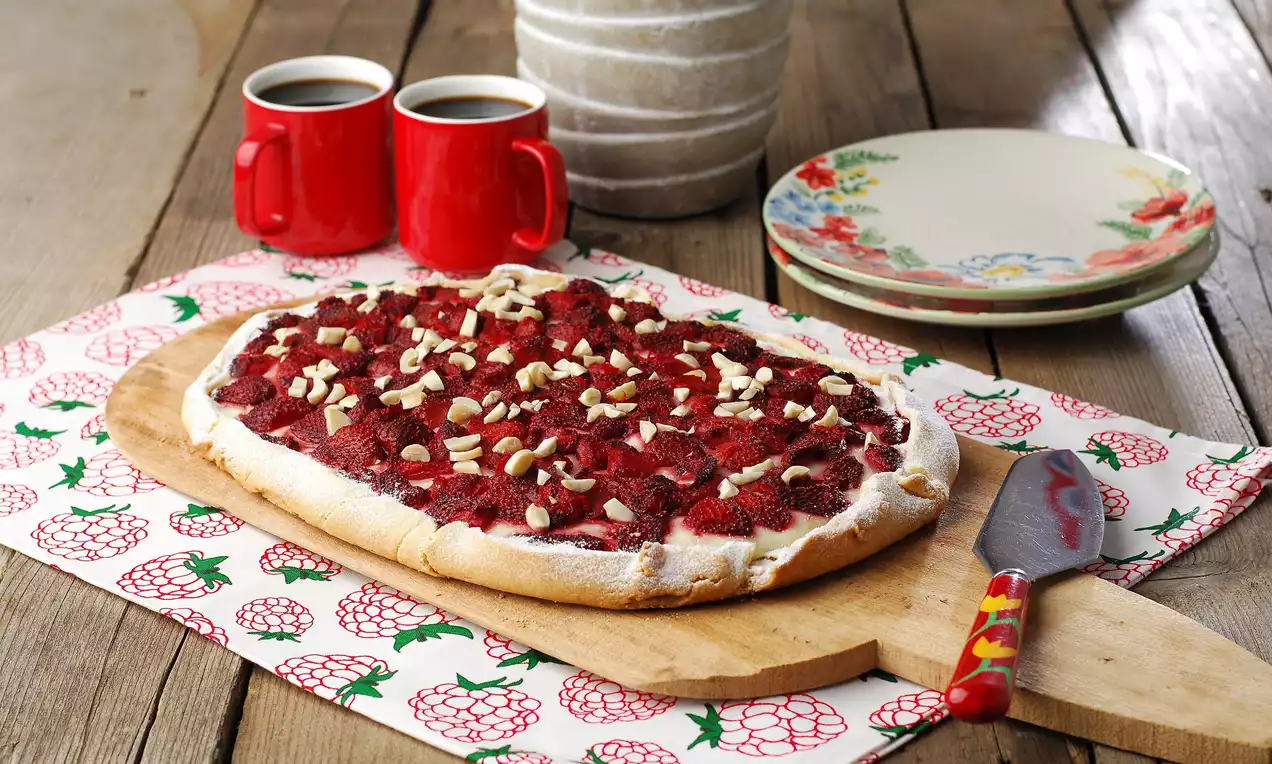 Ahşap bir servis tahtasında, beyaz pudra kaplı gevrek taban üzerinde taze çilekler ve serpilmiş badem parçalarıyla sunulmuş tart