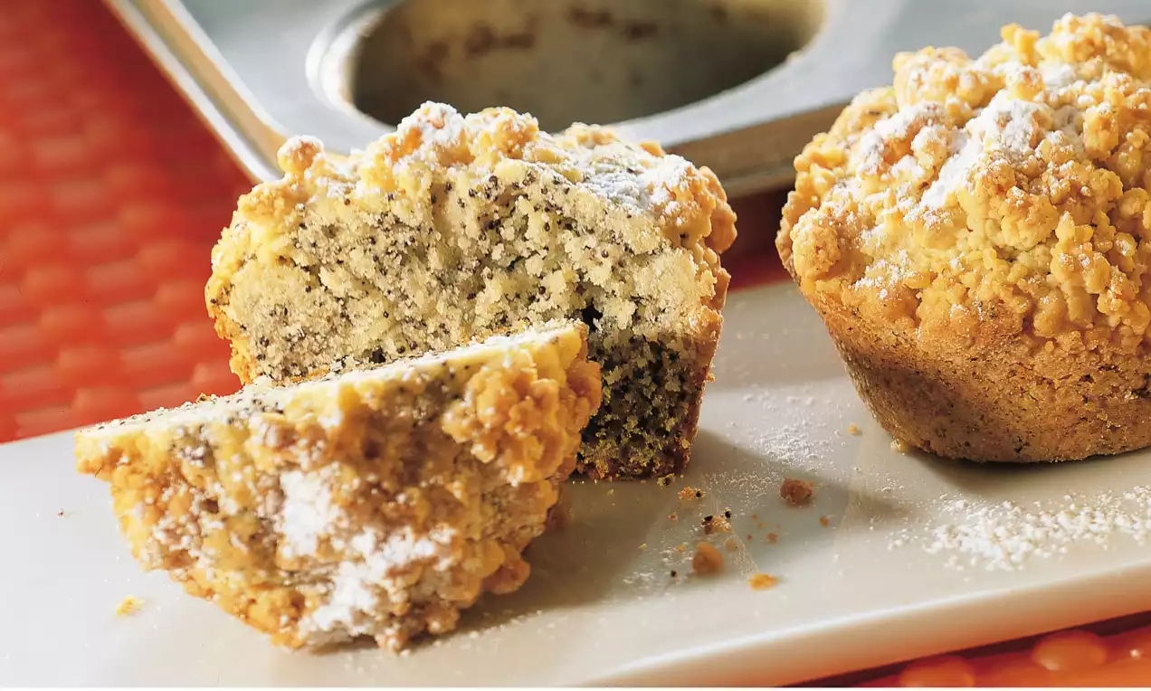 Saftige Mohn-Streusel-Muffins mit goldbrauner Kruste und Streuseln, ein Muffin halbiert auf weißer Platte vor einer Backform bestäubt mit Puderzucker