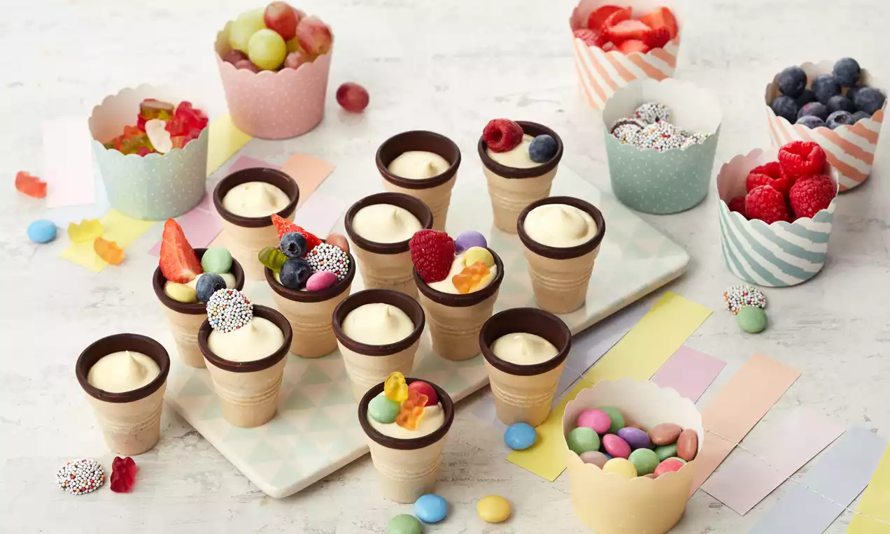 Kleine Dessert-Becher aus Waffel mit heller Creme, bunten Früchten und Schokodekoration, umgeben von farbigen Cupcake-Förmchen und Streudeko