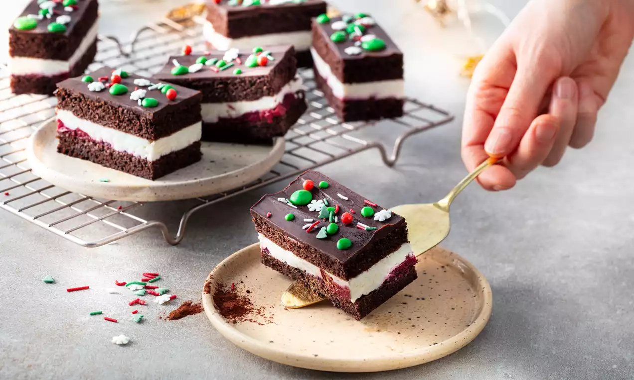 Donkere chocoladeslices met witte roomlaag, versierd met groene en rode sprinkles en M&M's op een beige bord en metalen rooster