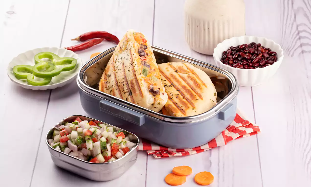 Golden brown grilled panini triangles stuffed with spiced rajma, served in a blue box with chopped salad, red chilies, and capsicum slices