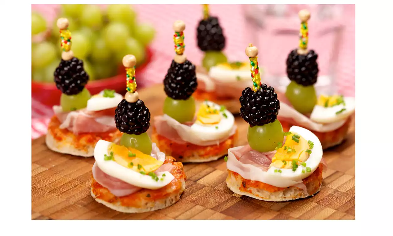 Kleine runde Brotstückchen mit Prosciutto, Ei, Schnittlauch und dekorativen Spießen aus Brombeeren und grünen Weintrauben auf Holzbrett