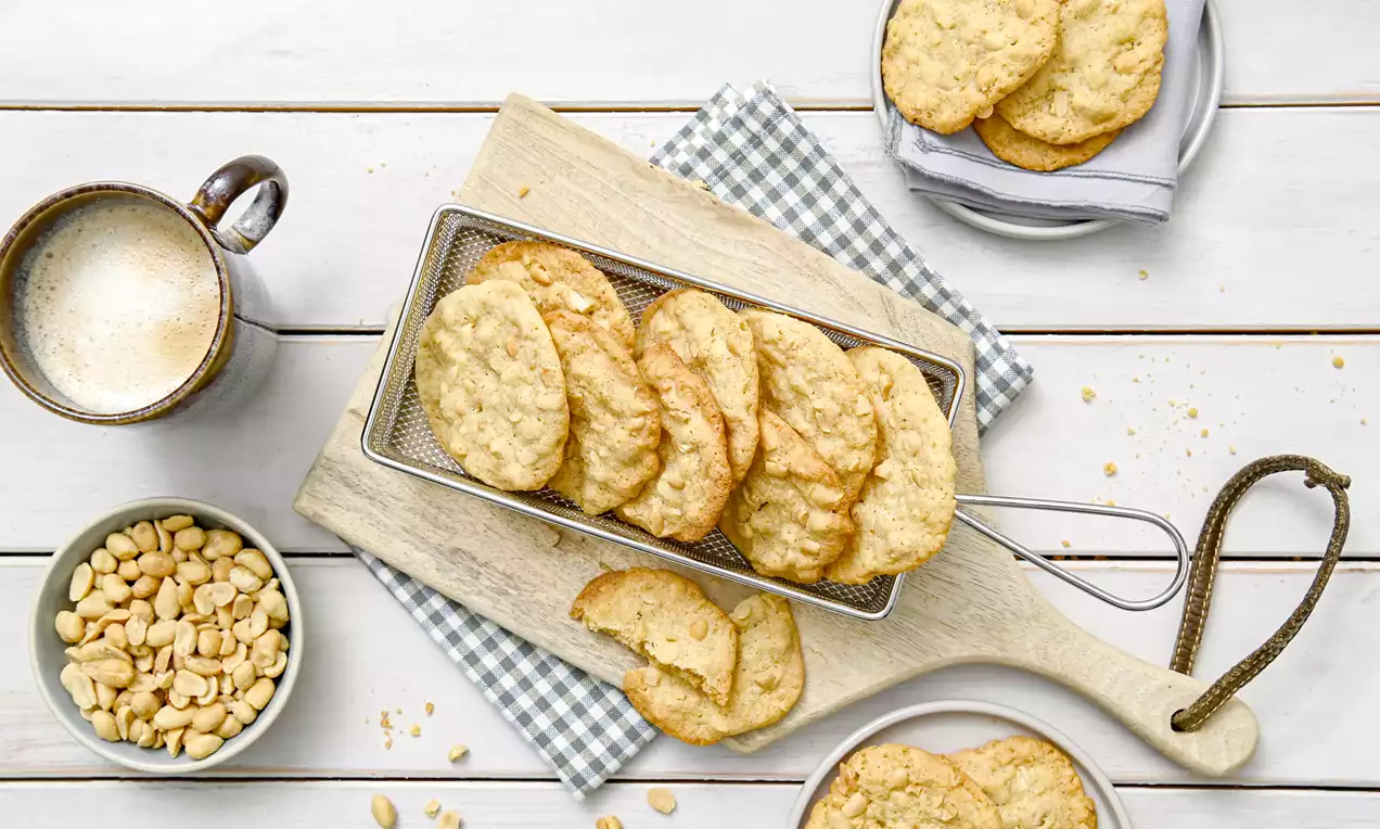 Erdnuss-Cookies goldbraun, rund und leicht gekrümmt mit feiner, leicht unregelmäßiger Oberfläche auf Holzbrett und silbernem Drahtkorb drapiert