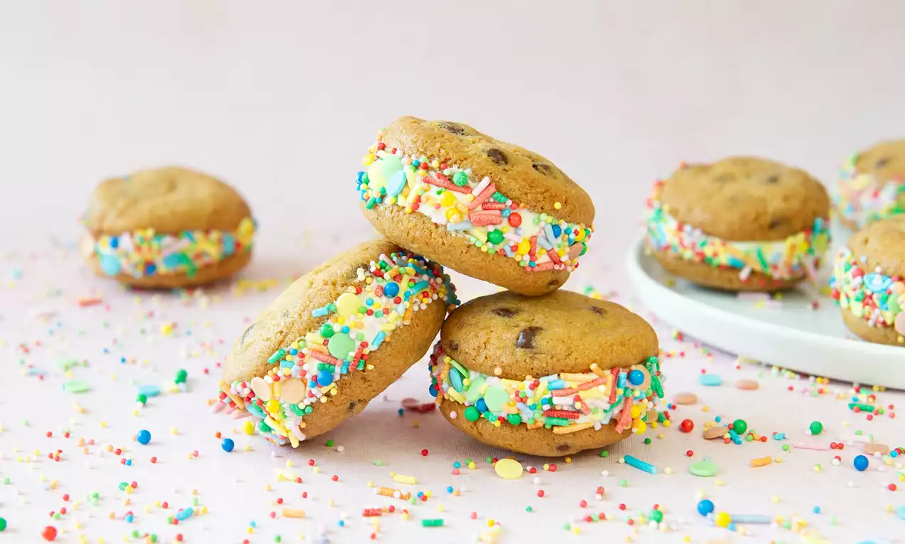 Bruine sandwichkoekjes gevuld met witte room, versierd met kleurrijke sprinkles, liggend op een lichtroze ondergrond met losse decoratie