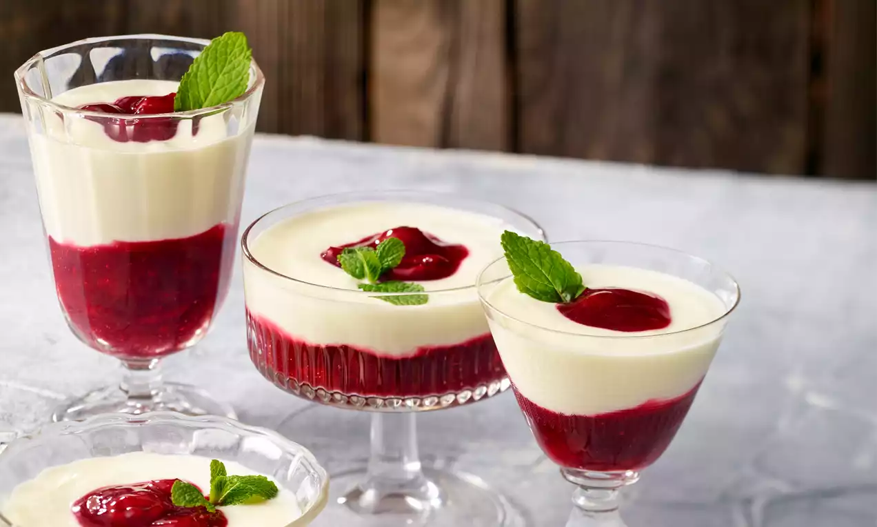 Topfen-Creme in klaren Gläsern mit weißer, glatter Oberfläche, auf roter Fruchtsoße, dekoriert mit frischen Minzblättern