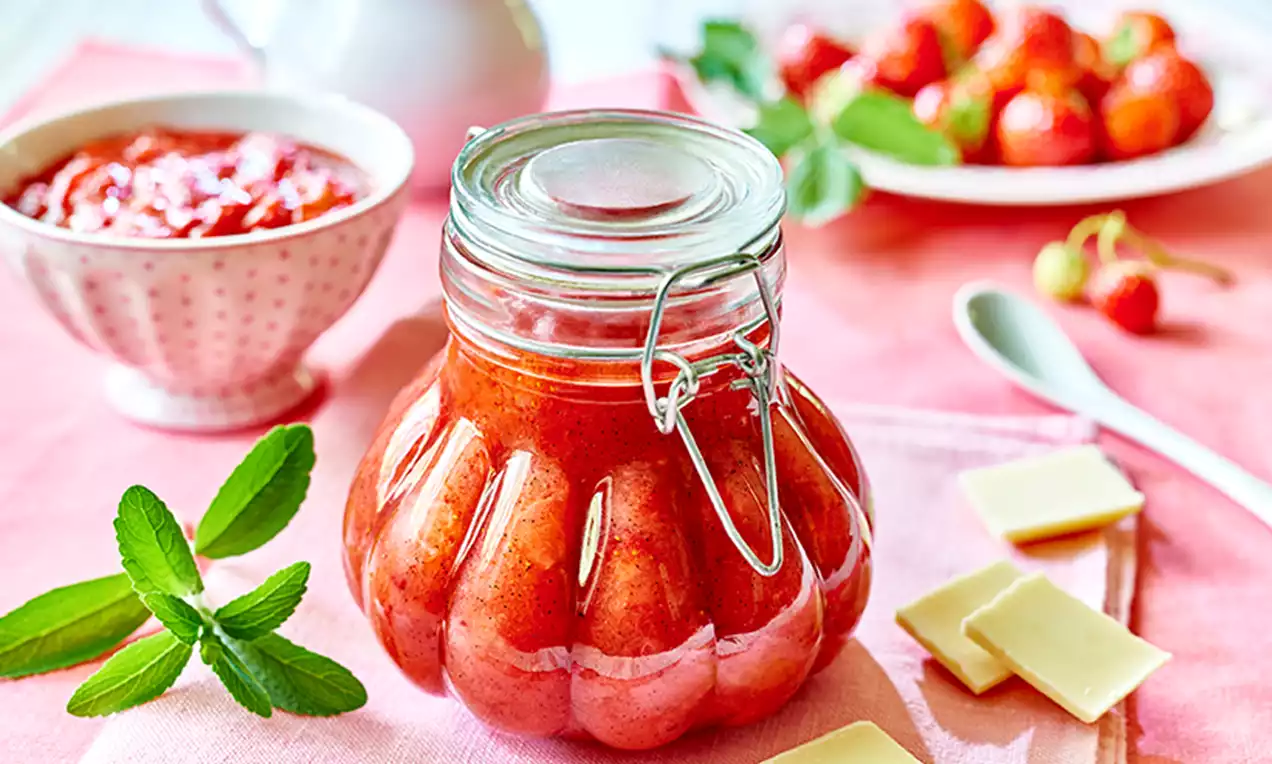 Słoik o falistej fakturze wypełniony truskawkowym dżemem o intensywnej barwie, otoczony miętą, kawałkami białej czekolady i świeżymi truskawkami