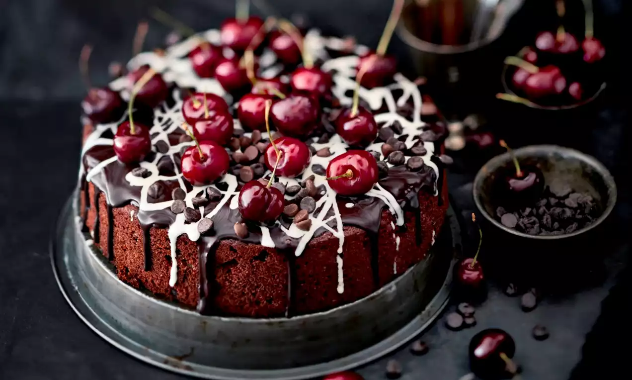 Korkea suklaatäytekakku tumman kiiltävällä suklaakuorrutteella, valkoisilla suklaaraitoina valuneilla koristuksilla ja punaisia kirsikoita päällä