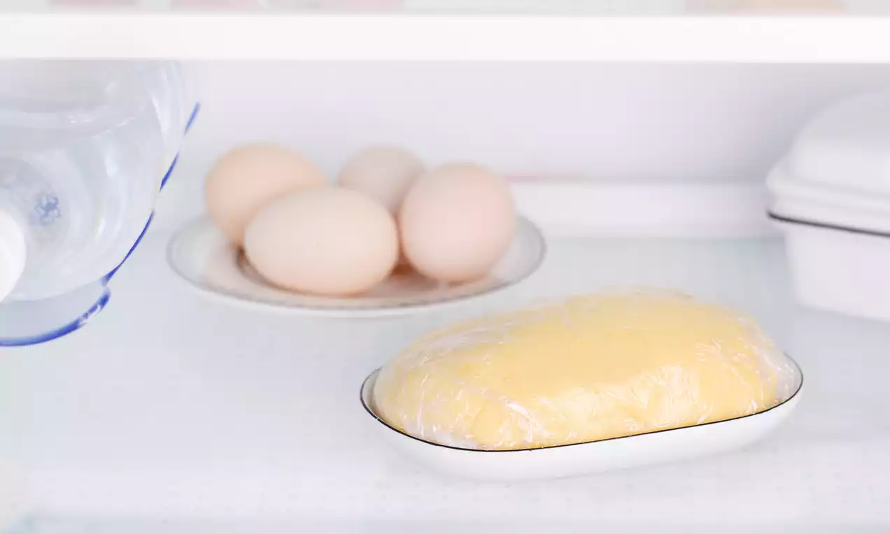 Kruche ciasto o złocistym kolorze, owinięte w przezroczystą folię, spoczywa na białym talerzyku obok jajek na porcelanowym półmisku