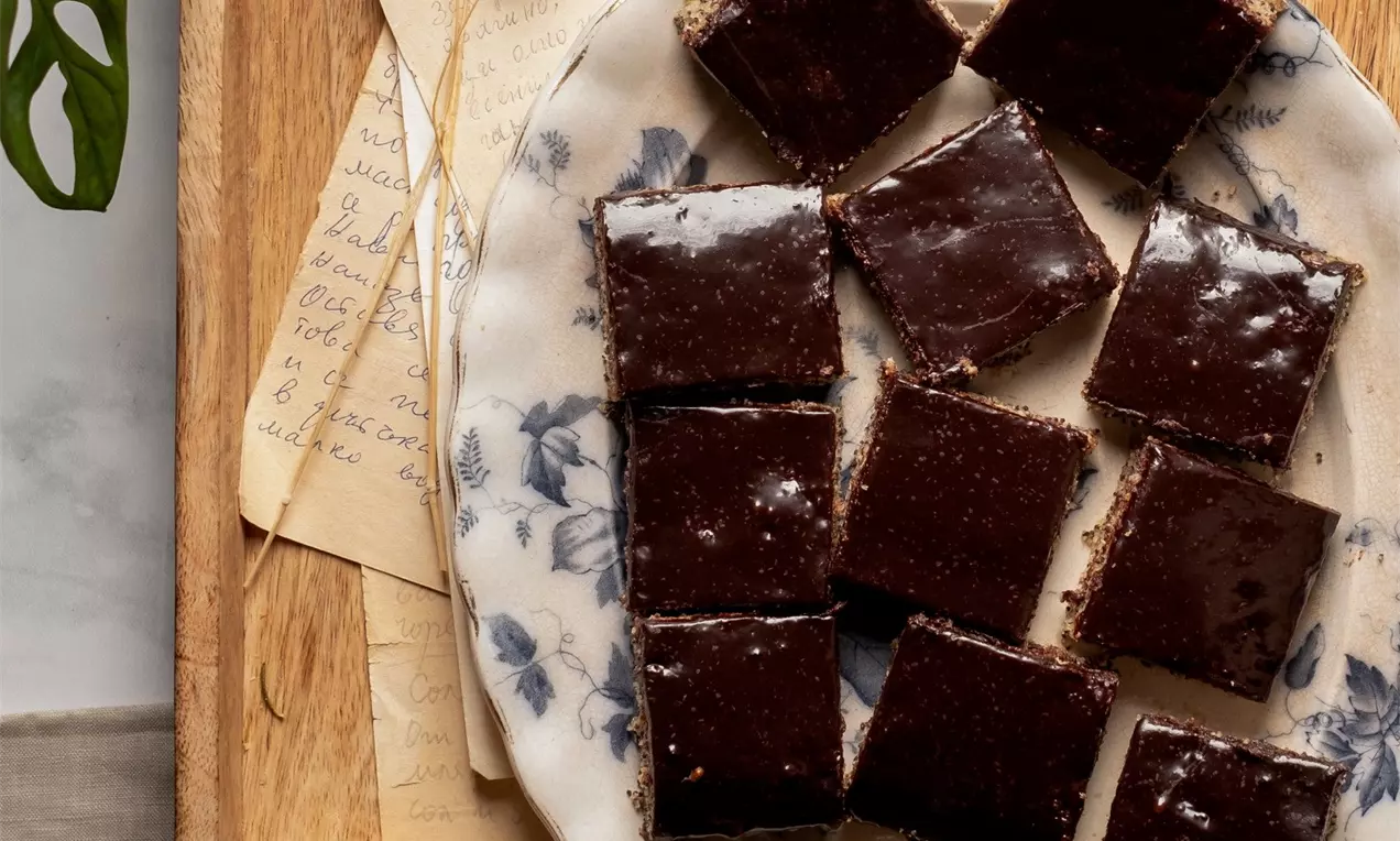 Квадратни парчета маков сладкиш с лъскава шоколадова глазура, разположени равномерно върху бяла чиния със сини флорални мотиви