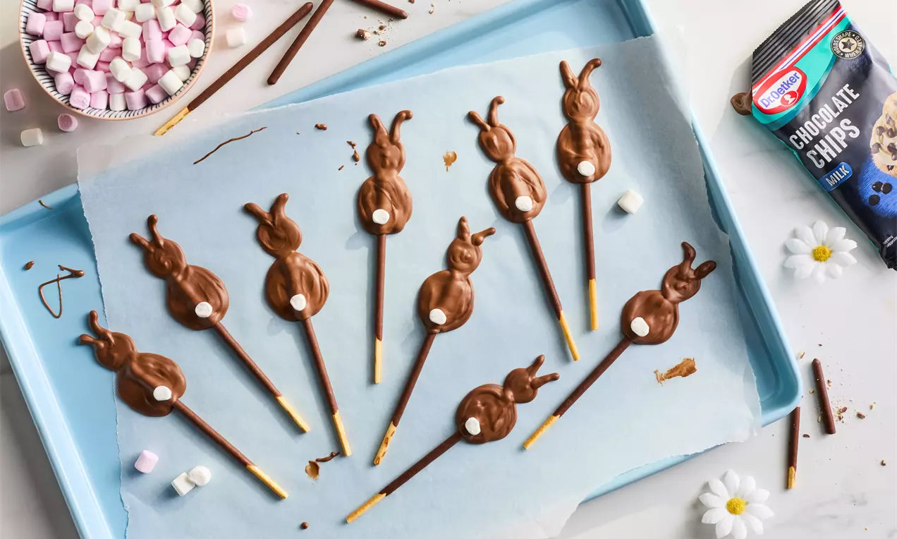 Milk chocolate bunny-shaped pops with pretzel stick handles, topped with white chocolate dots, arranged on parchment paper on a blue tray
