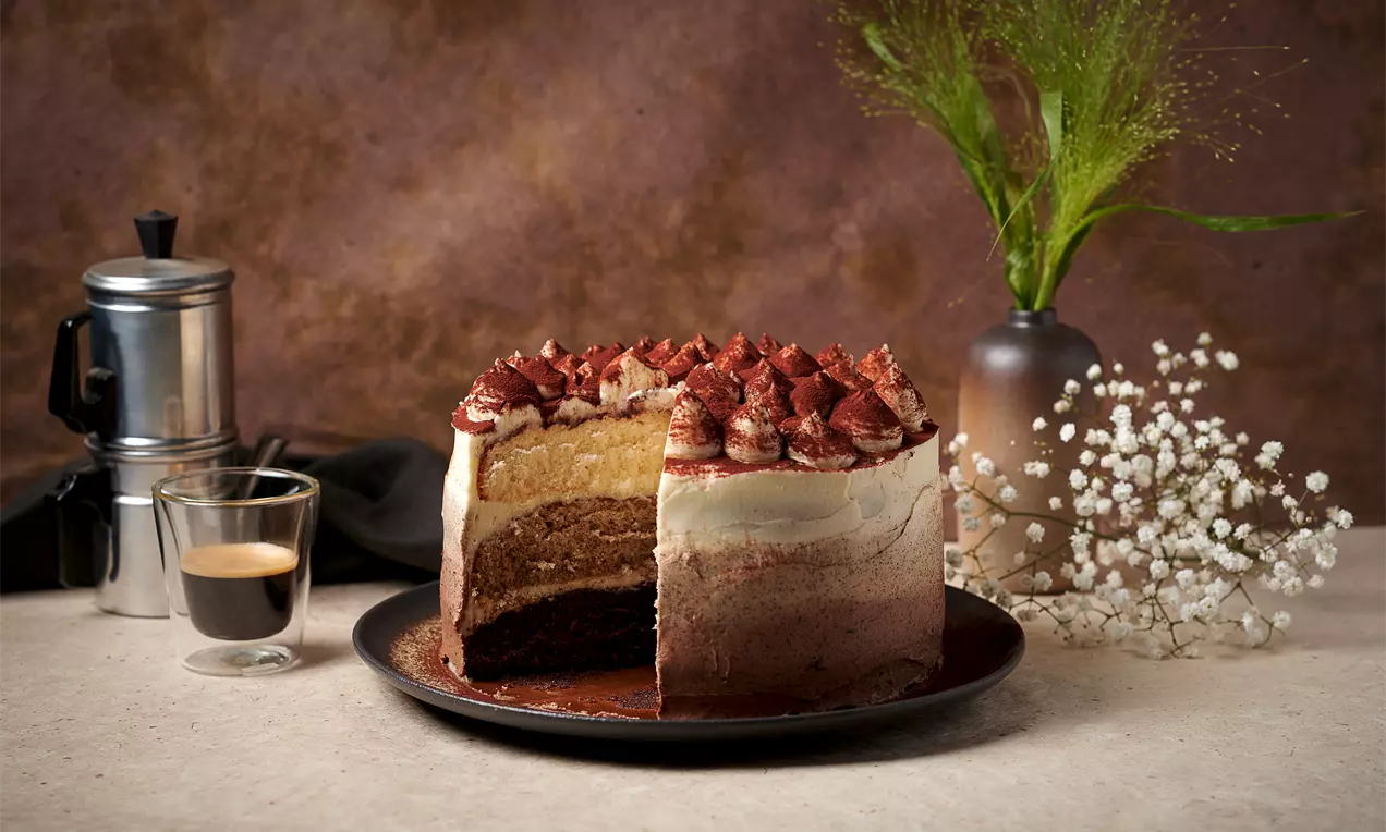 Многослойна омбре торта с плавен преход от тъмнокафяв до кремав цвят, покрита с какао и крем, върху тъмна чиния с небрежни декорации