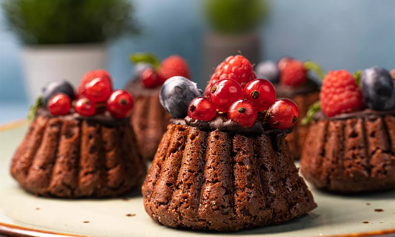 Кафяво-гладки мини кексчета с набраздена текстура, гарнирани с лъскави червени и сини горски плодове върху зелена чиния